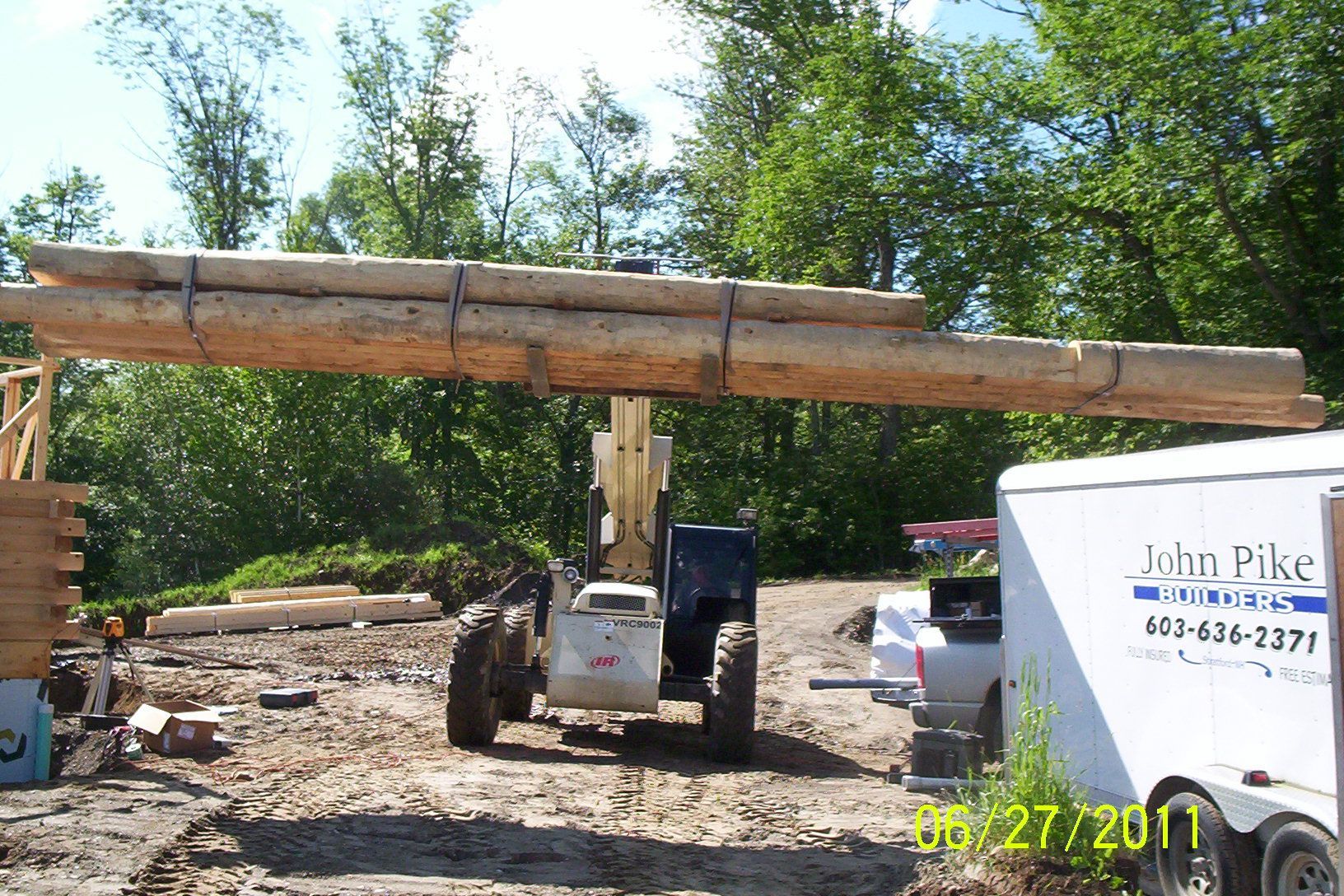 Telehandler lifting a long log beam on a wooded construction site with a John Pike truck nearby