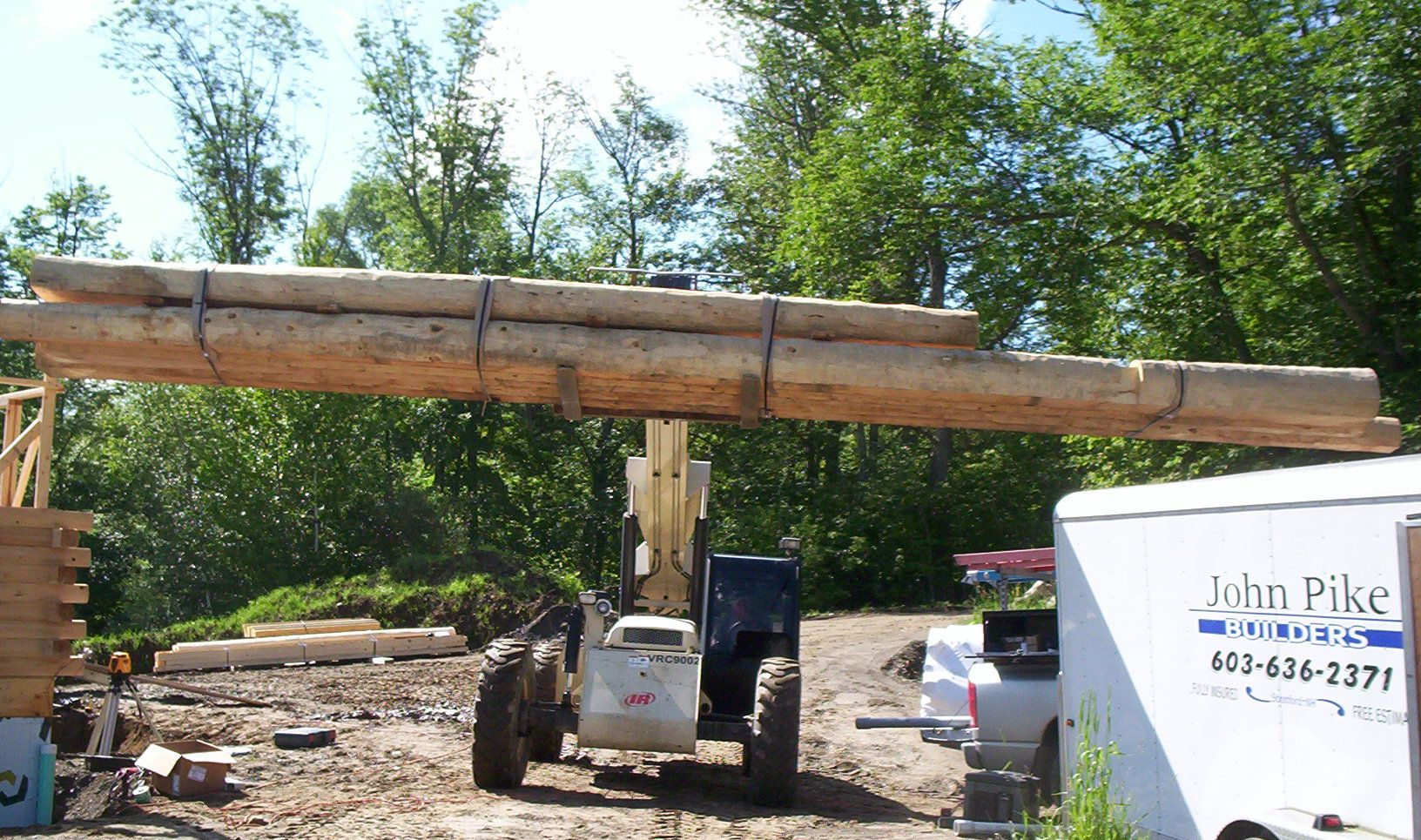 Construction scene with a lifted wooden beam, a skid steer, and a truck near a white John Pike sign.