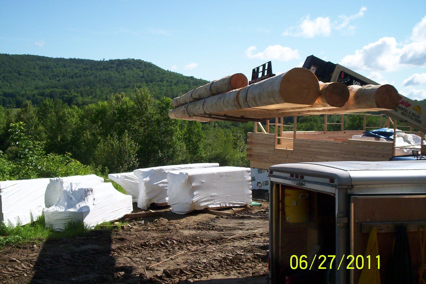 Logging truck unloading logs at a construction site with wrapped materials and wooded hills in the background