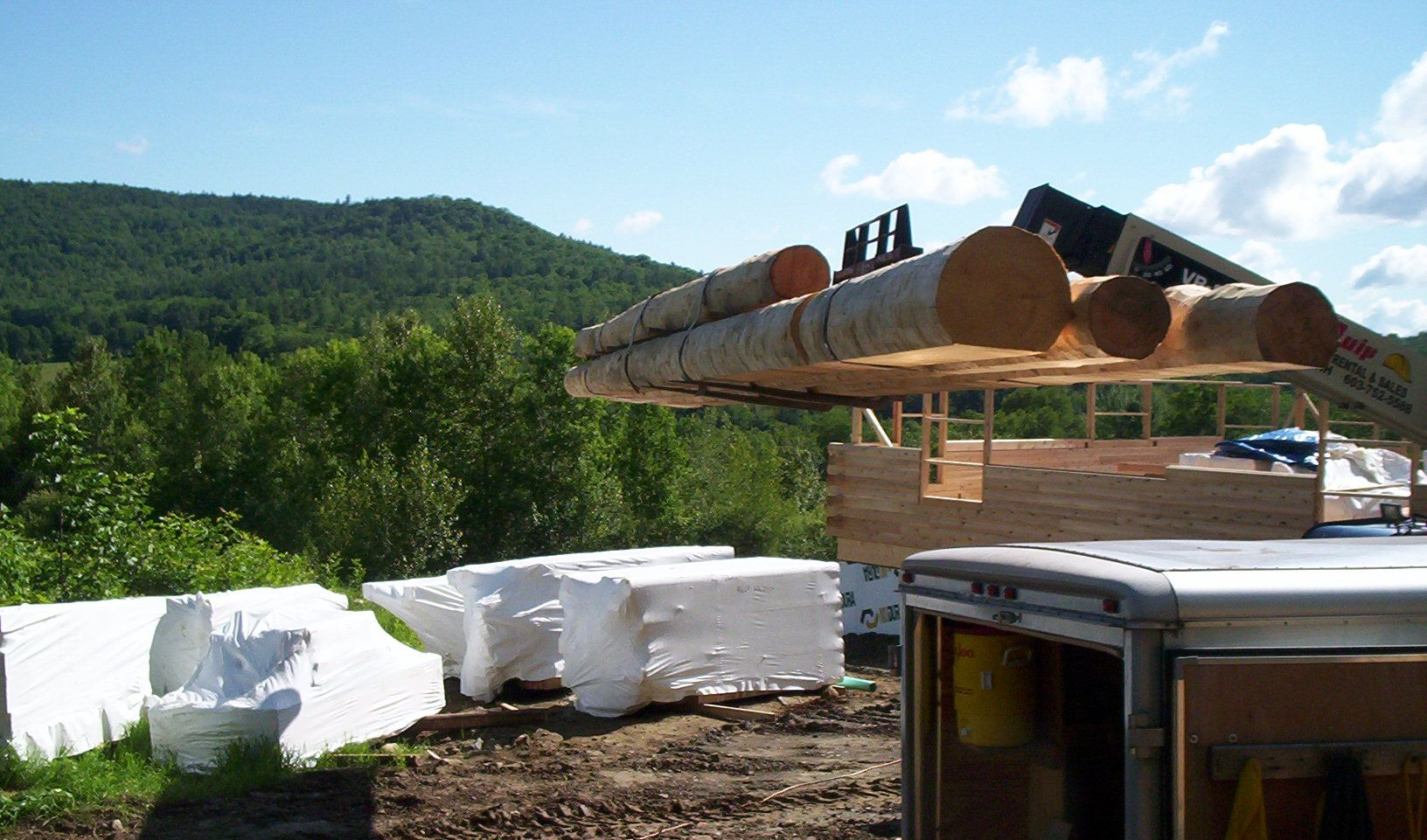 Large log suspended by crane above white-wrapped materials at a forested construction site