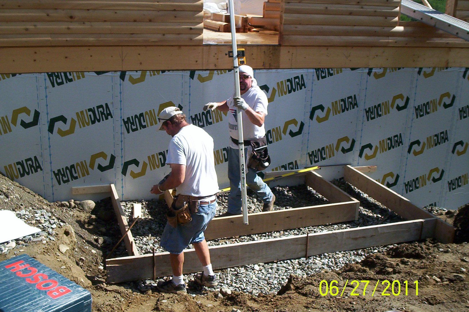 Two workers framing a house foundation beside a wall wrapped in weather barrier