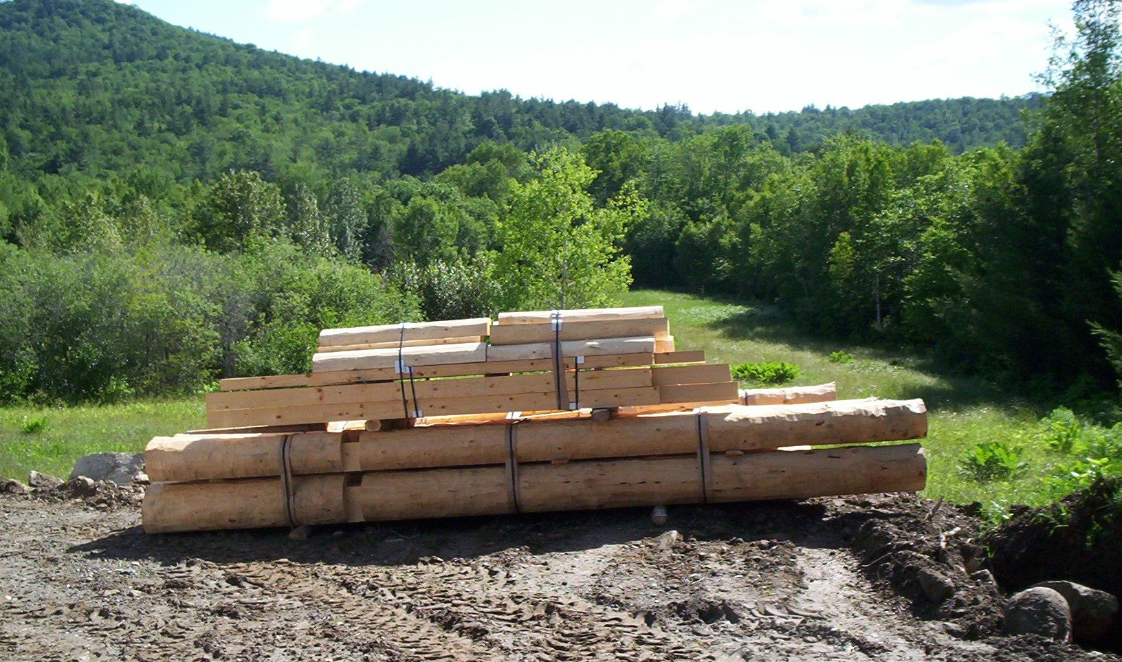 Stacked lumber in a clearing with green forested hills in the background.