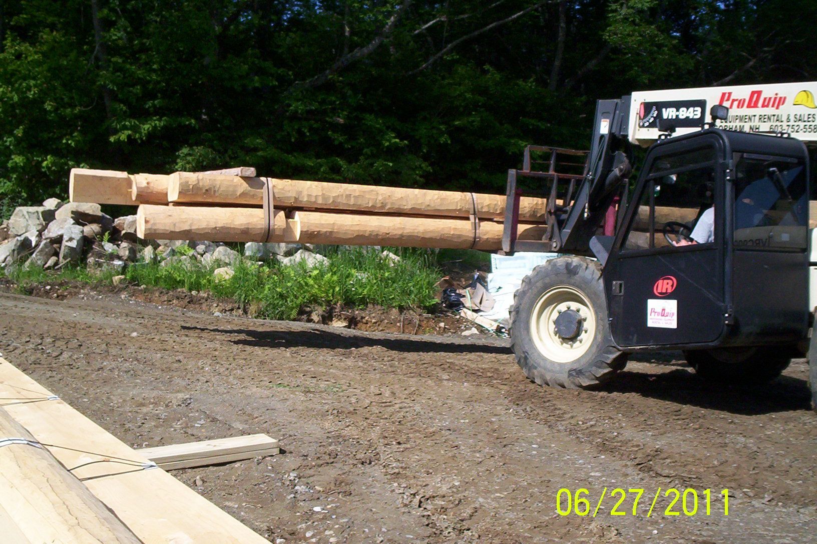 Logs loaded on a flatbed truck beside a gravel lot and wooded area.
