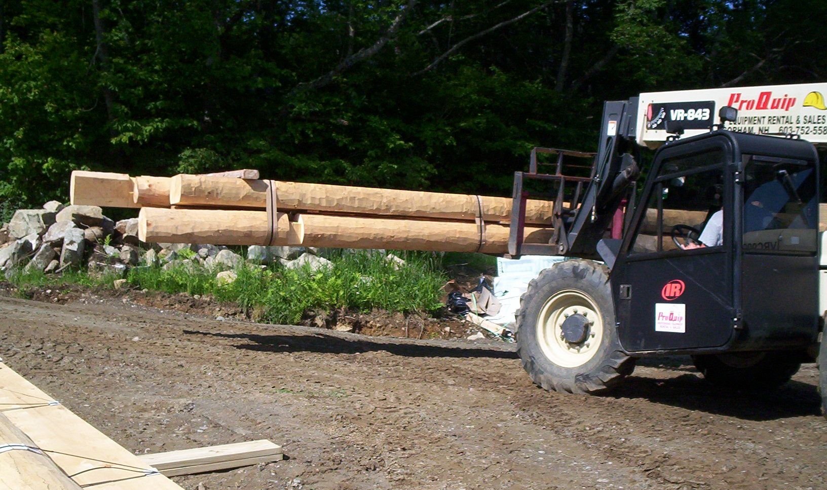 Forklift carrying logs on a gravel lot beside a wooded area