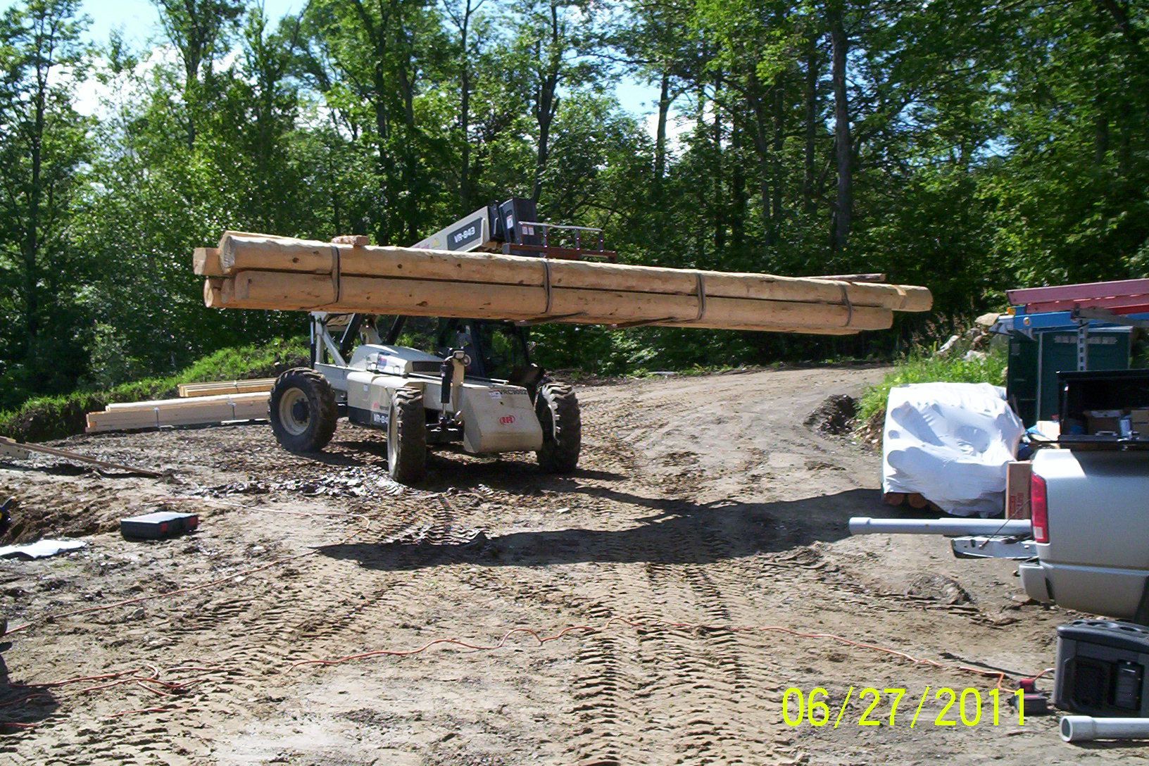 Construction vehicle carrying long lumber across a muddy site, with trees and equipment nearby.