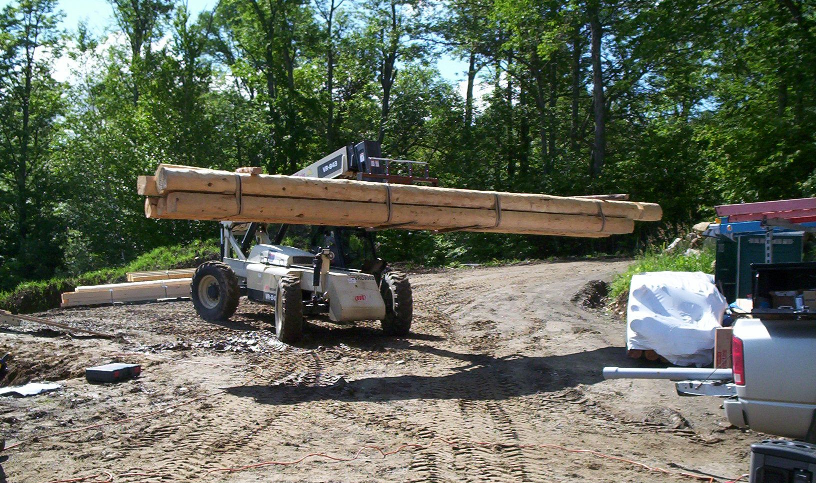 Forklift carrying long wooden beams across a dirt worksite with trees in the background