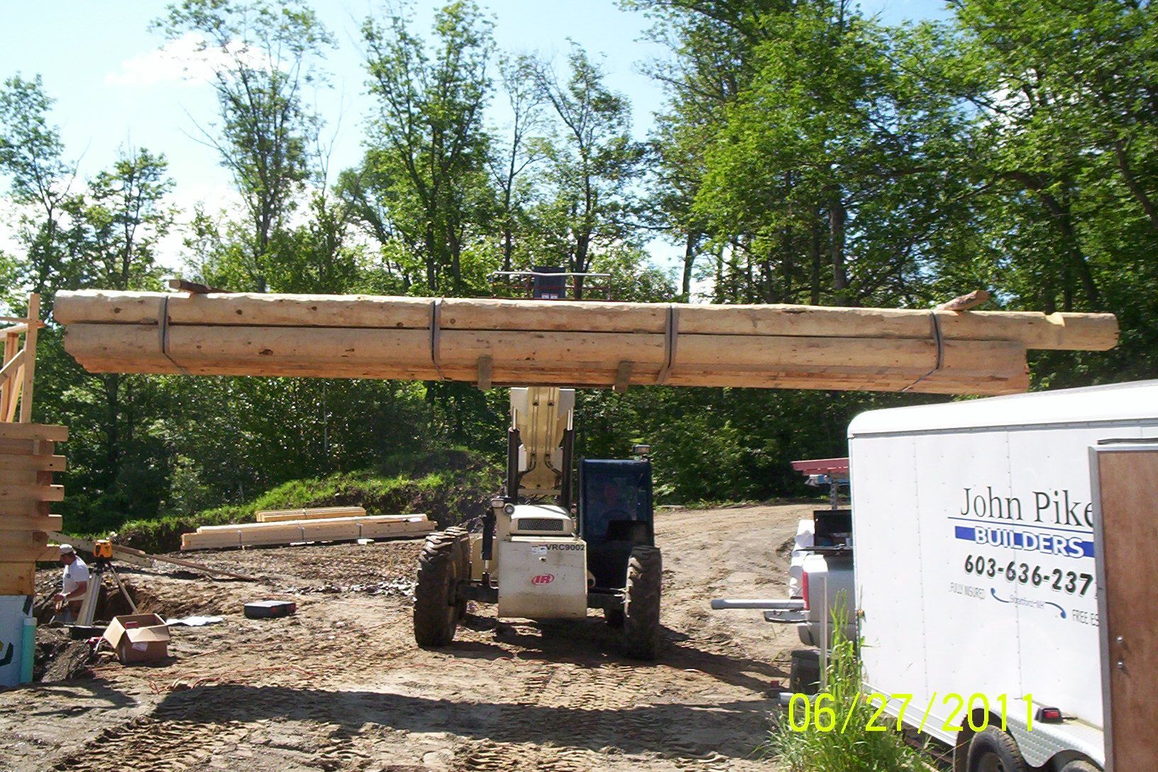 Forklift carrying a long wooden beam on a gravel lot, with trees and a white trailer nearby.