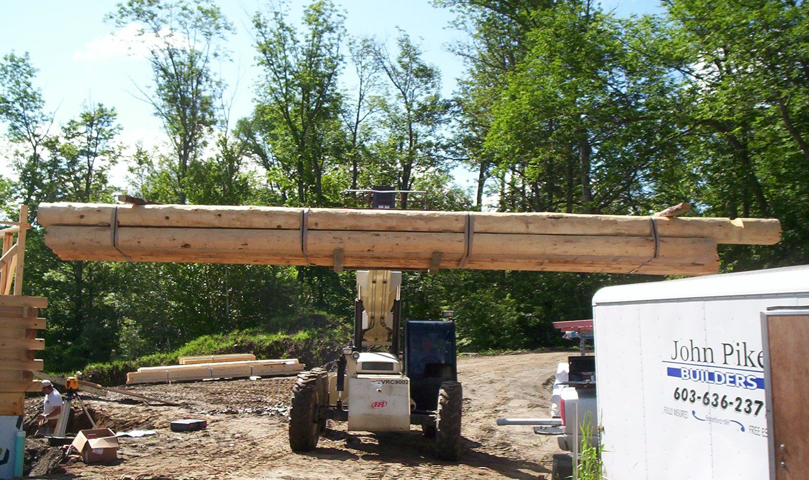 Skid steer lifting a long wooden beam on a construction site with trees in the background.