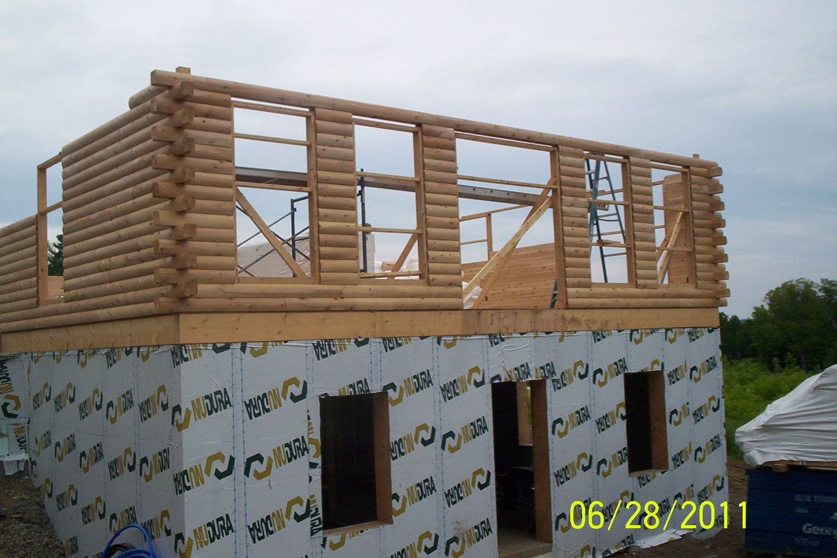 Partially built log-cabin style house with exposed upper framing and white wrapped lower walls