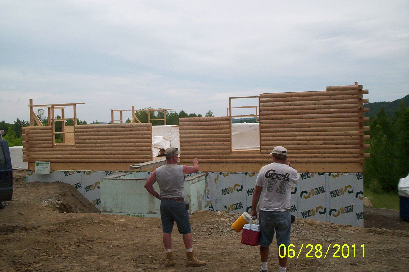 Two workers inspect a partially framed wooden wall at a construction site, with dirt piles and a date stamp.