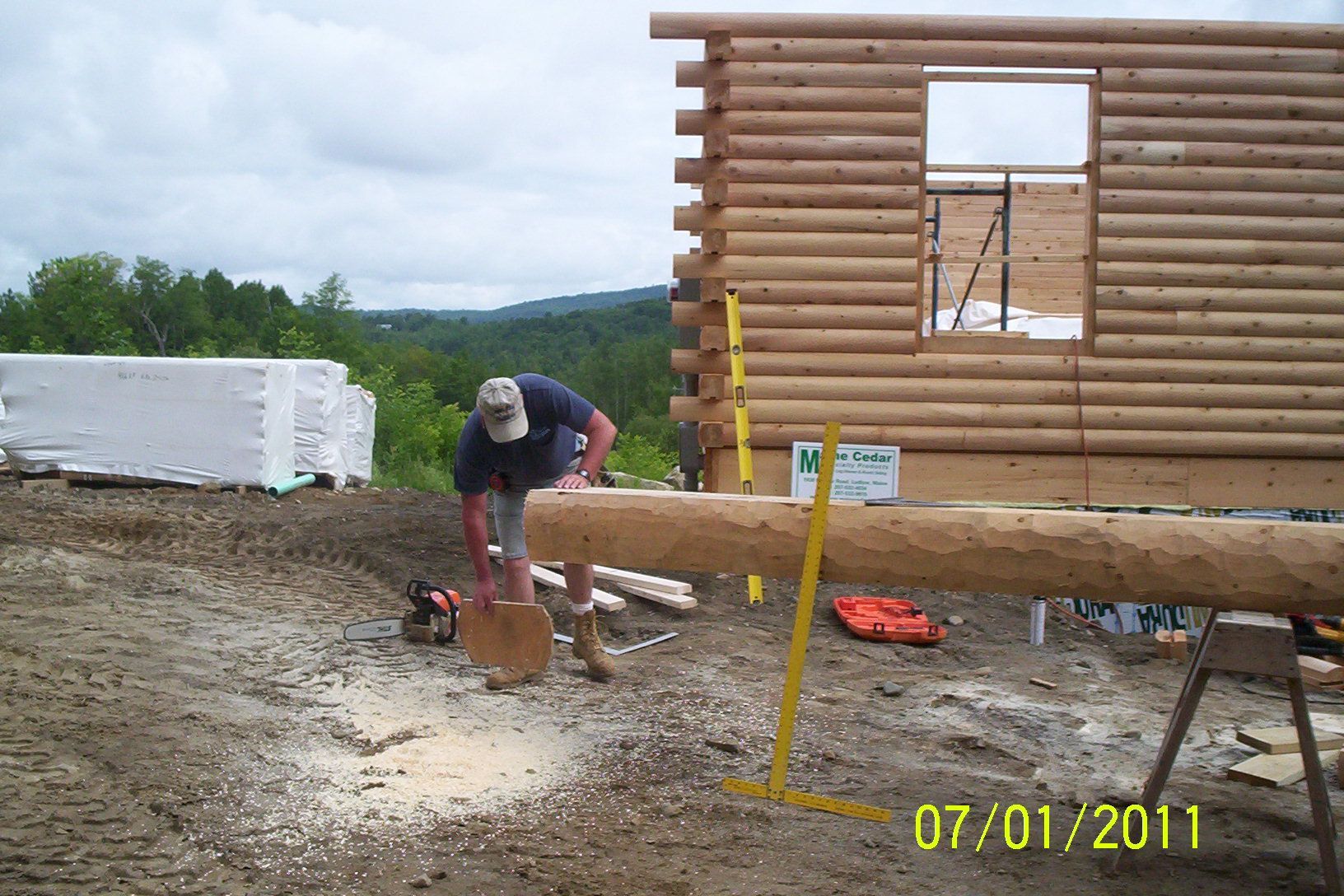 Person using a chainsaw near a log cabin under construction, with logs and building materials around.