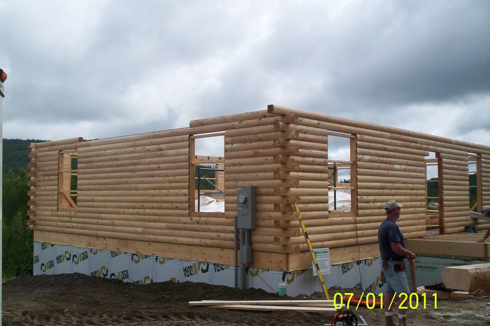 Partially built log cabin with window openings; a worker stands beside it under cloudy skies
