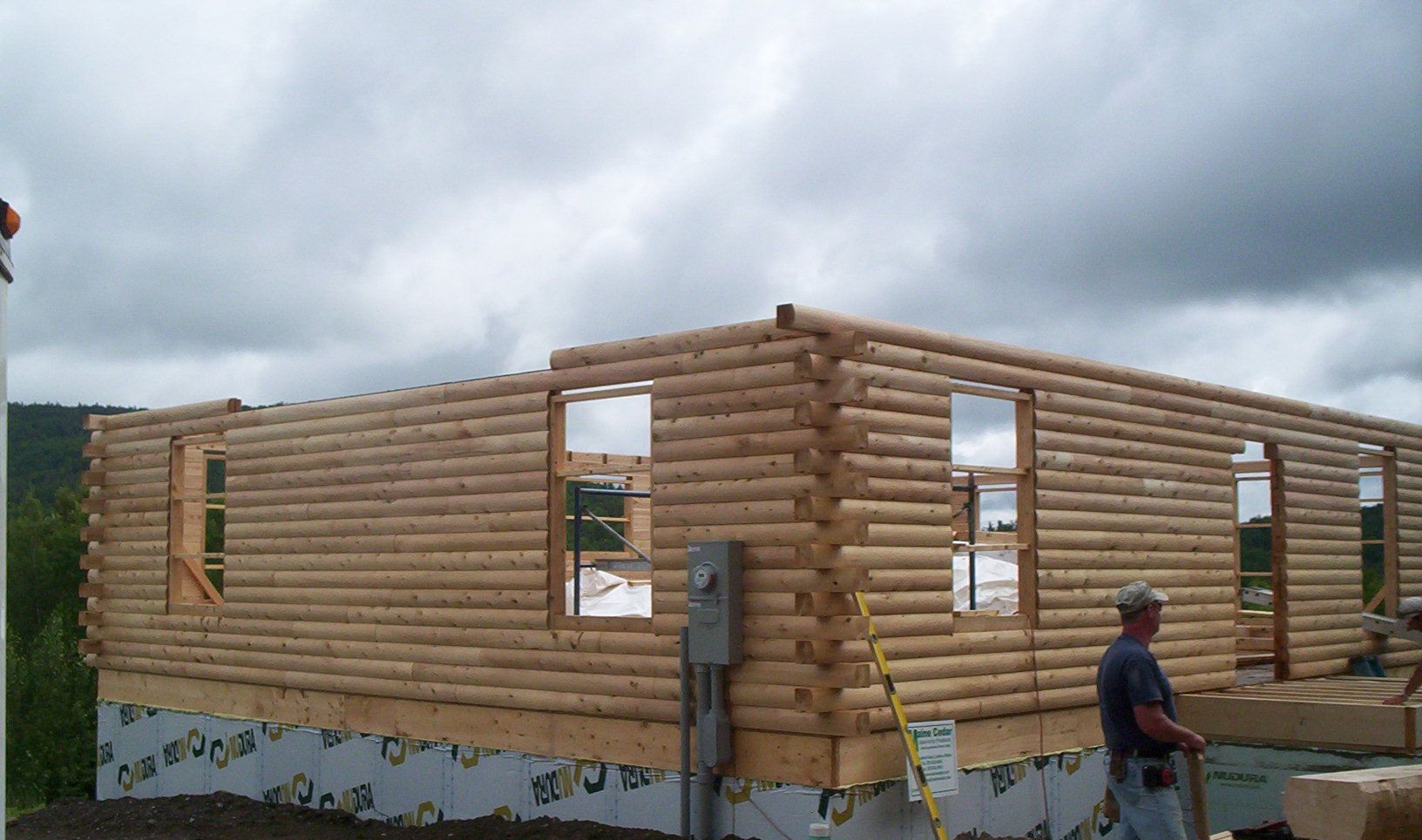 Partially built log cabin with open window frames under a cloudy sky, with a person standing nearby