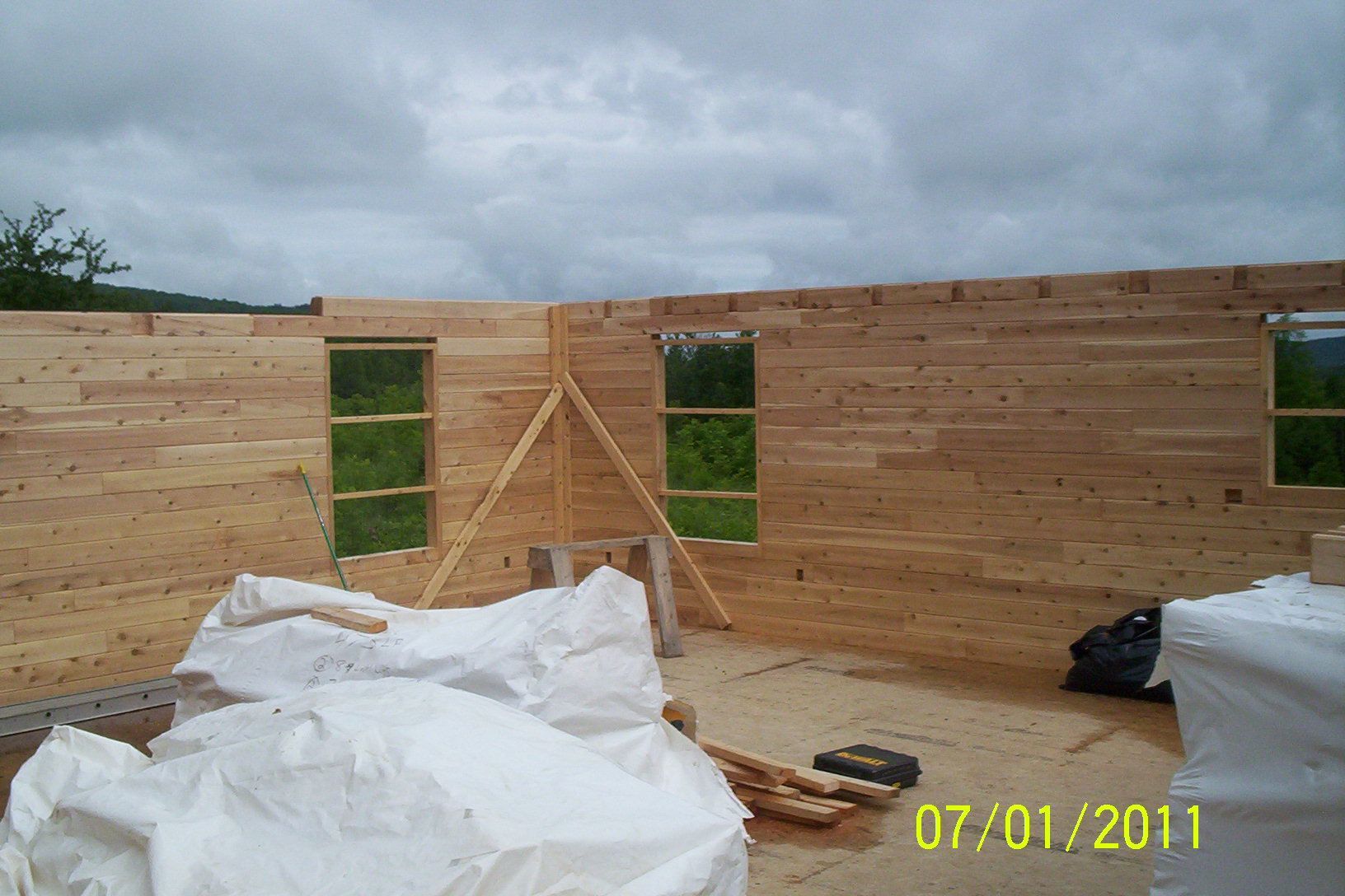 Partially built wooden house walls with window openings under a cloudy sky, construction materials in foreground