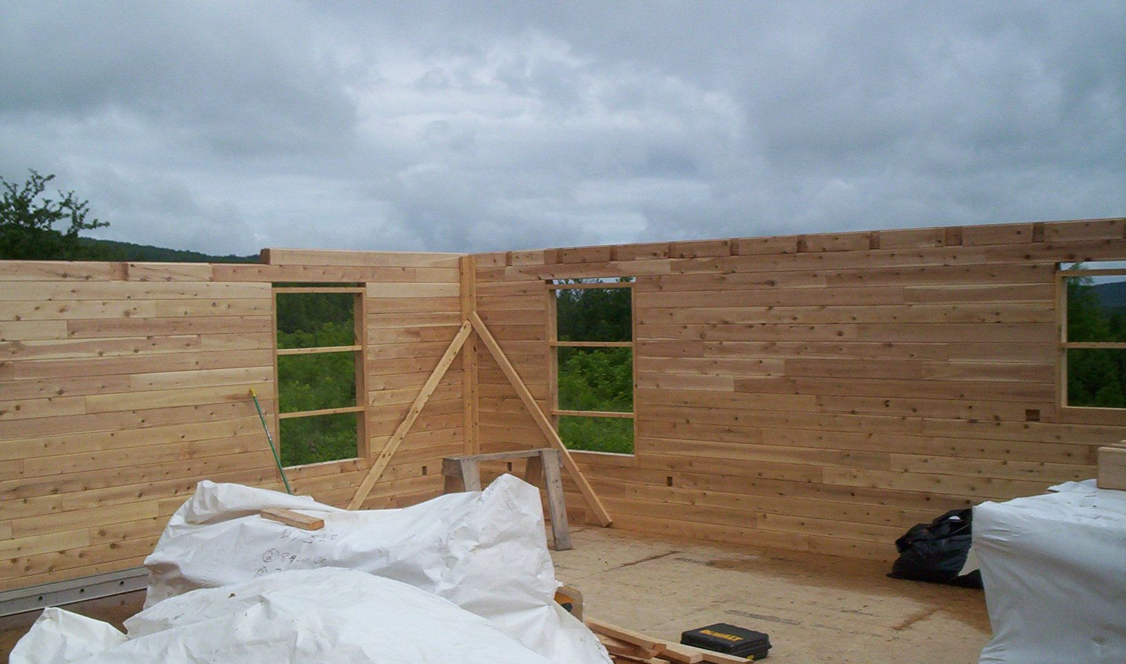 Partially built wooden structure with window openings and white bags in the foreground under a cloudy sky