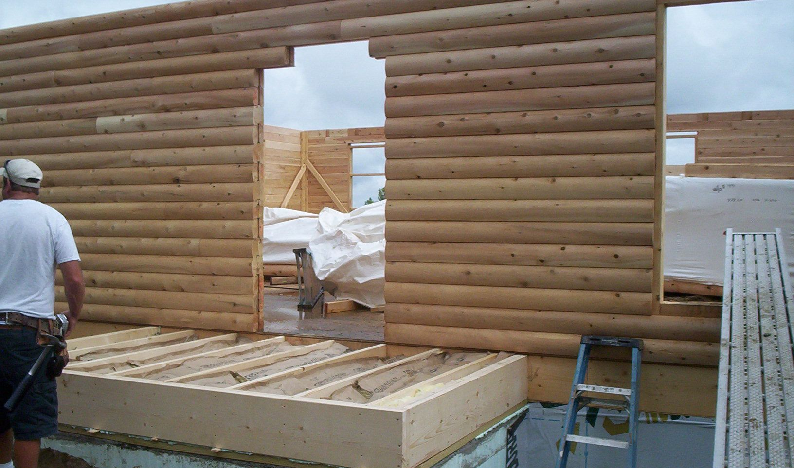 Worker building a log cabin wall with window openings and roof framing in progress