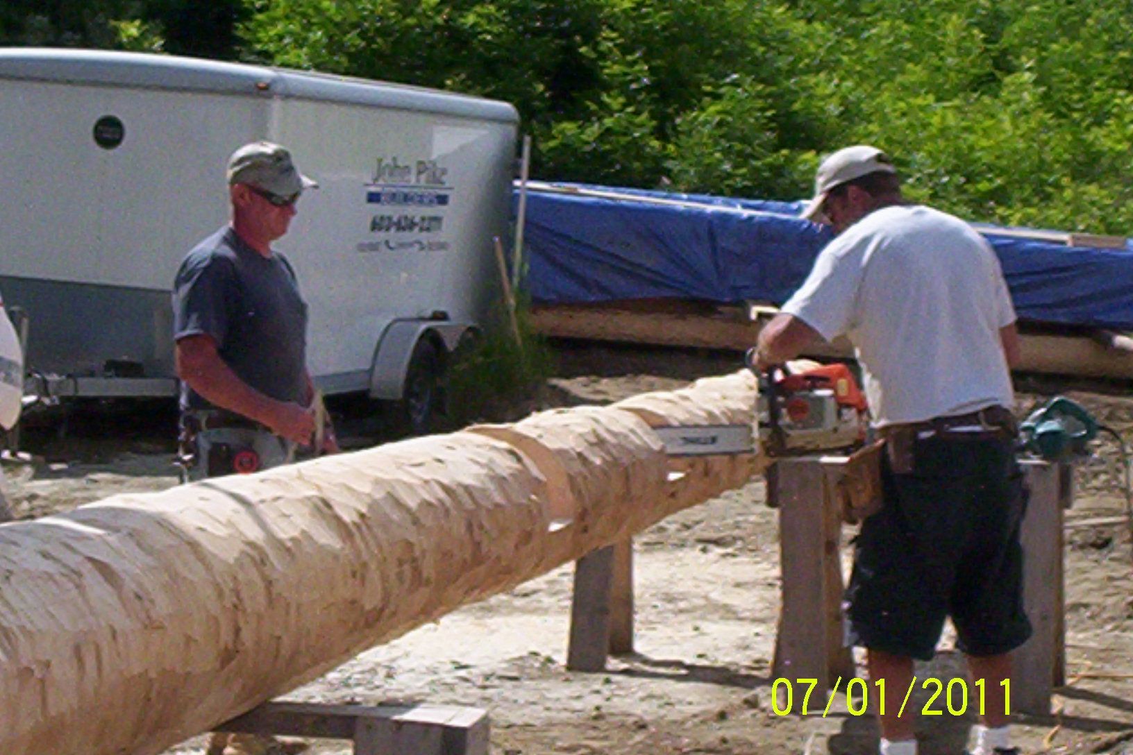 Two men work on a large peeled log with a chainsaw in an outdoor yard, near a trailer.