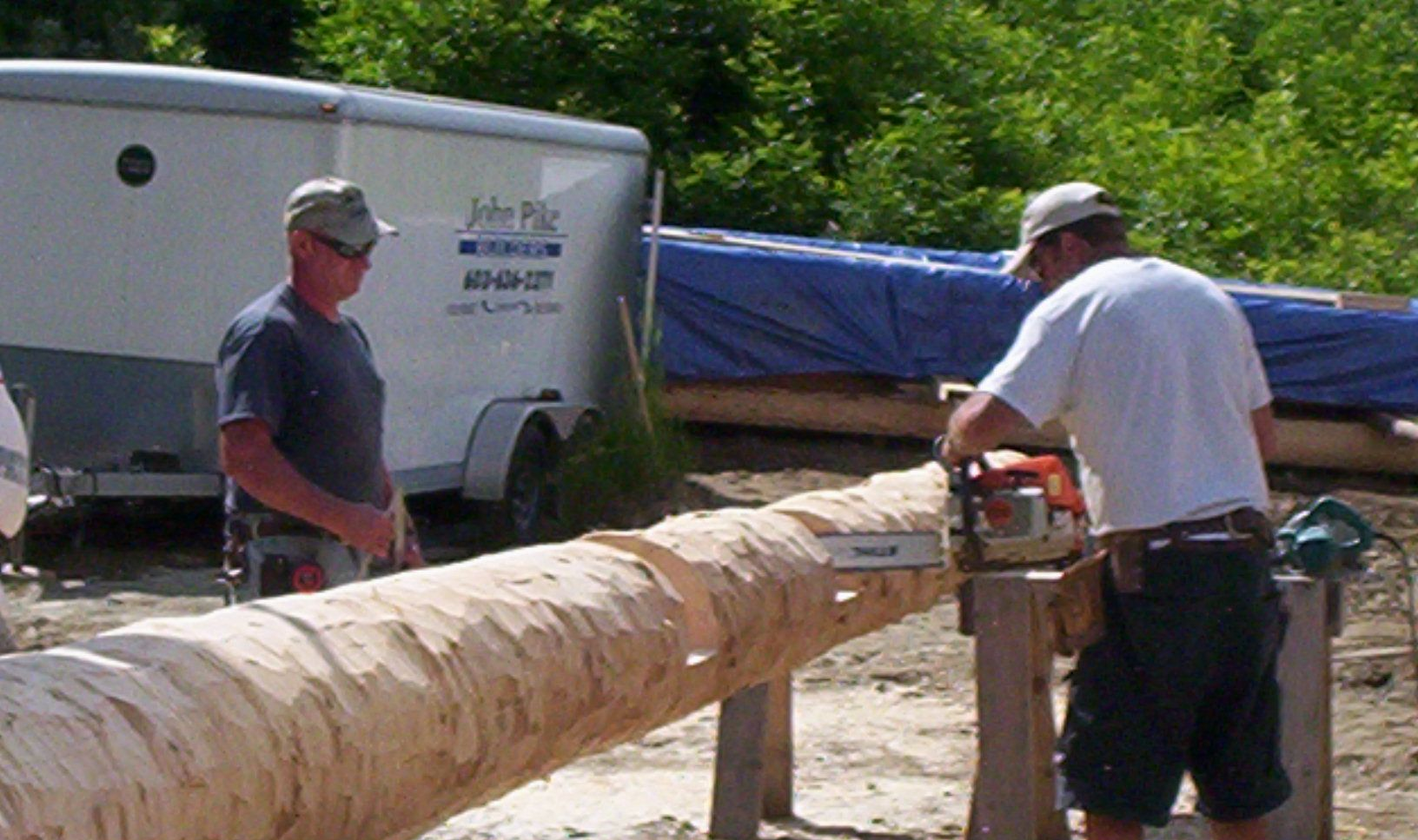 Two workers carving a large log outdoors with a chainsaw near a trailer