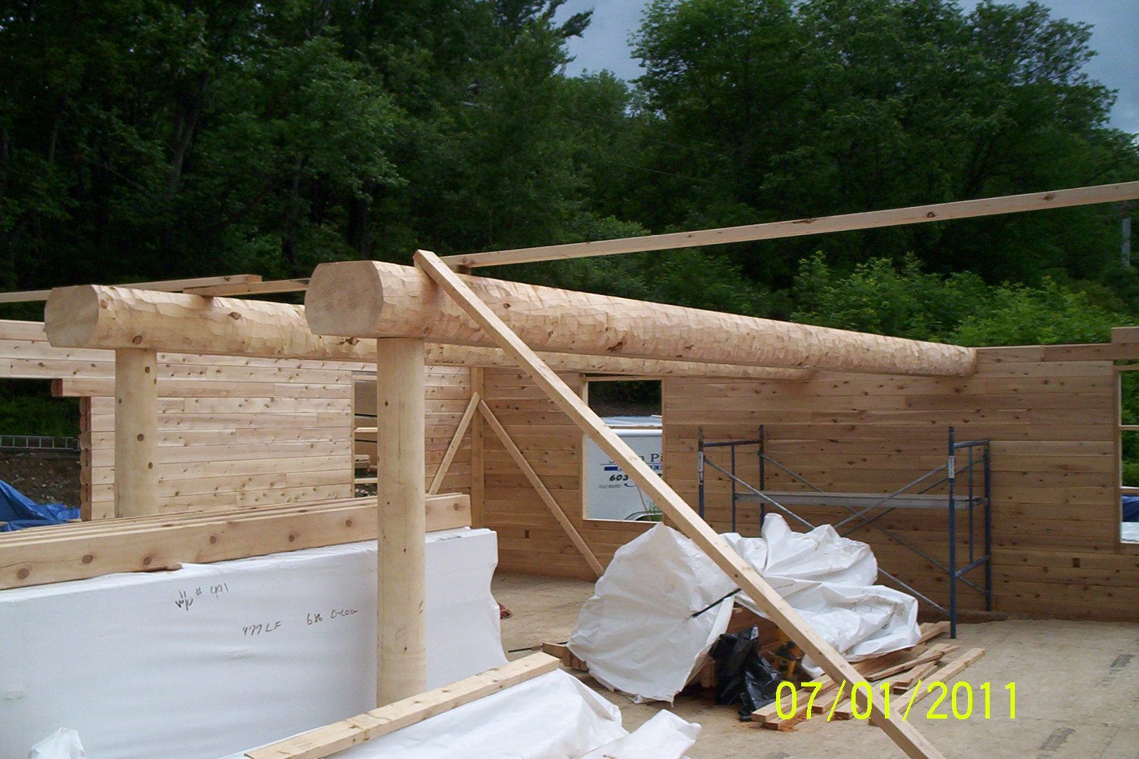 Partially built wooden house with log roof beams and white tarps at a construction site