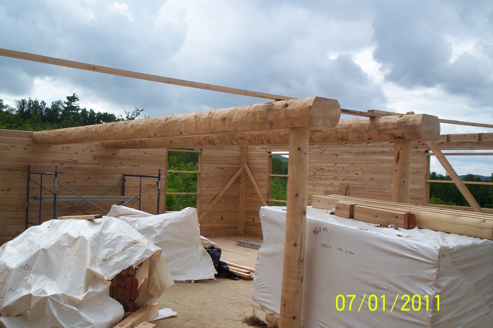 Wooden house frame under construction with tarps and scaffolding beneath a cloudy sky