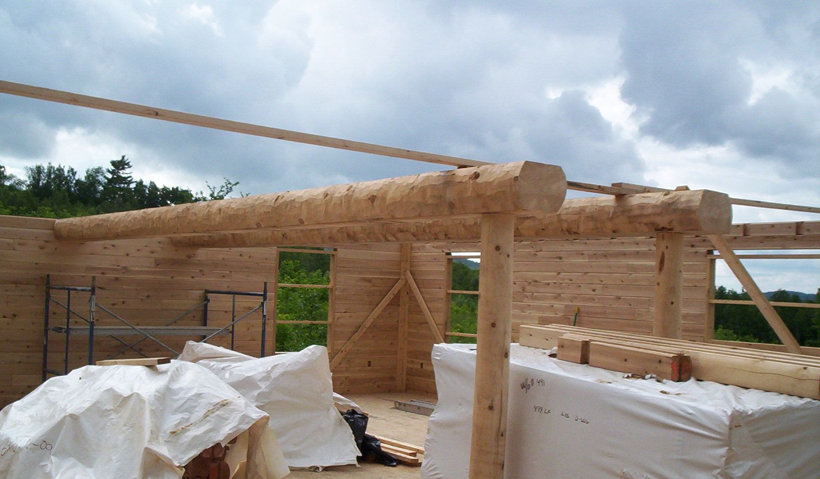 Wooden house under construction with roof beams, wrapped materials, and cloudy sky overhead