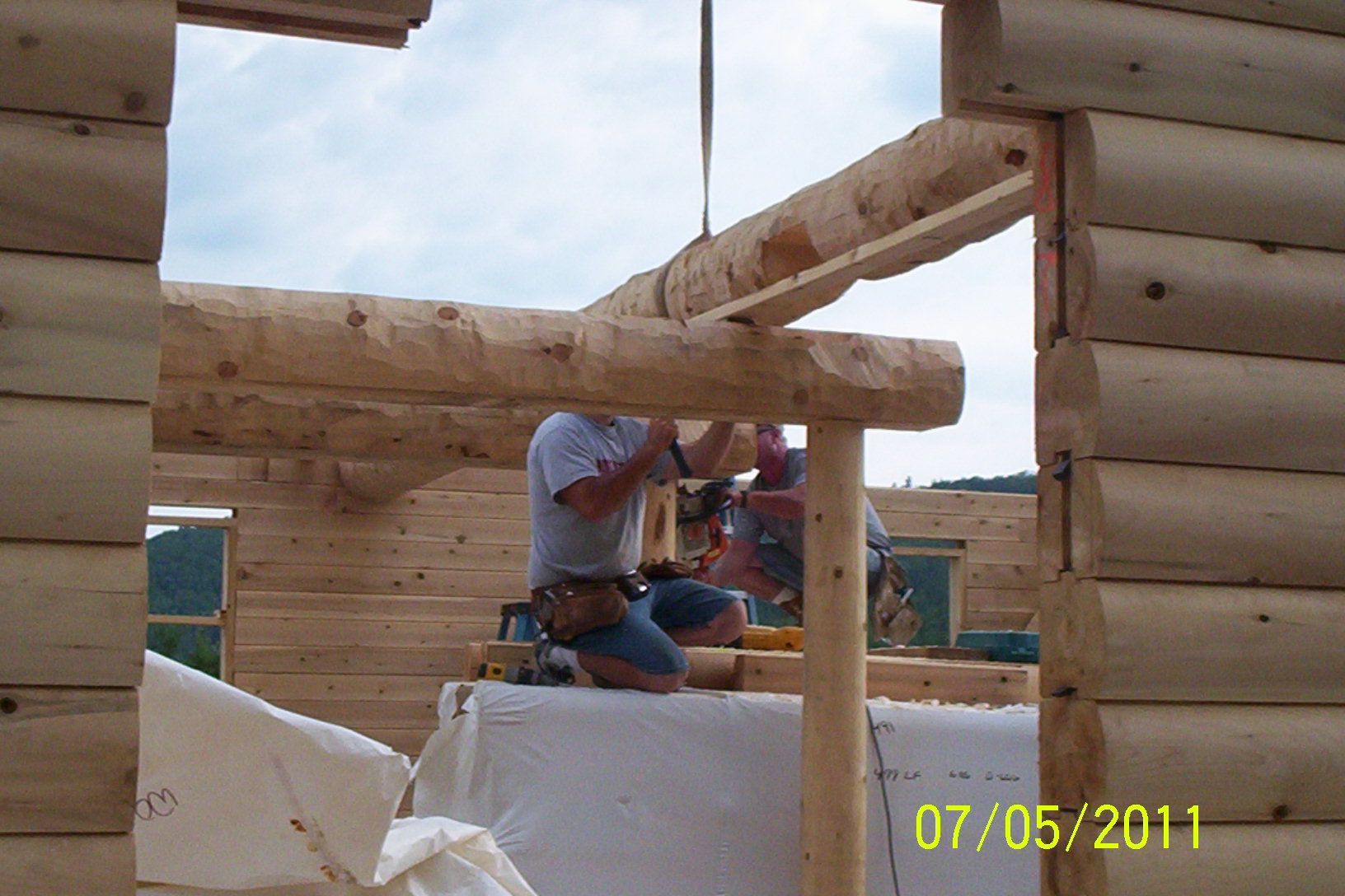 Worker installing wooden beams in a log cabin frame, with insulation and date stamp visible.