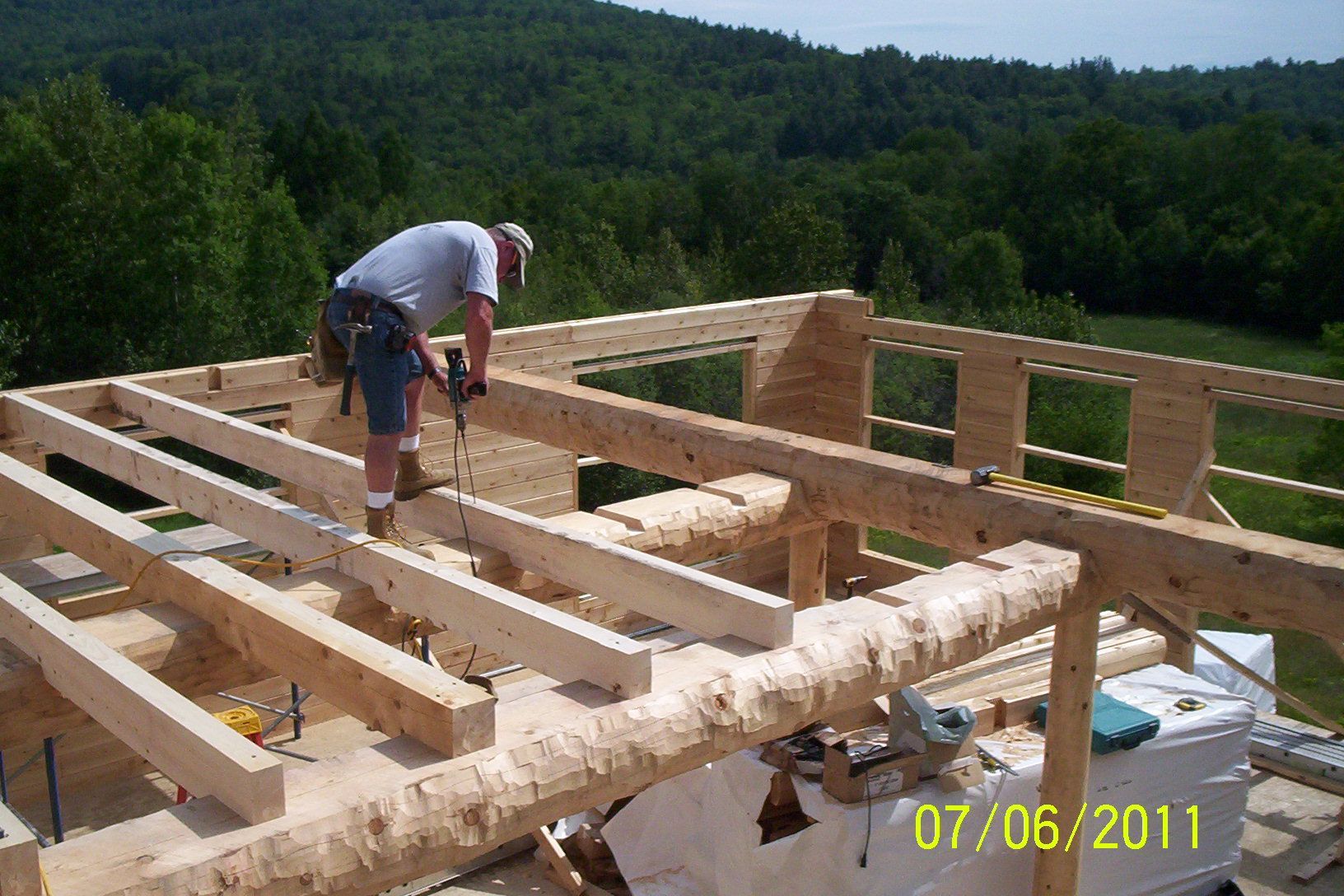 Worker assembling a wooden house frame on a hillside with forest in the background.