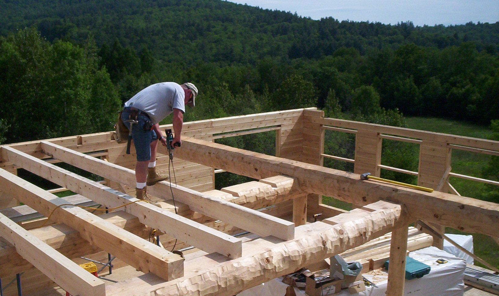 Worker building a wooden house frame on a hillside, using a hammer amid exposed beams.