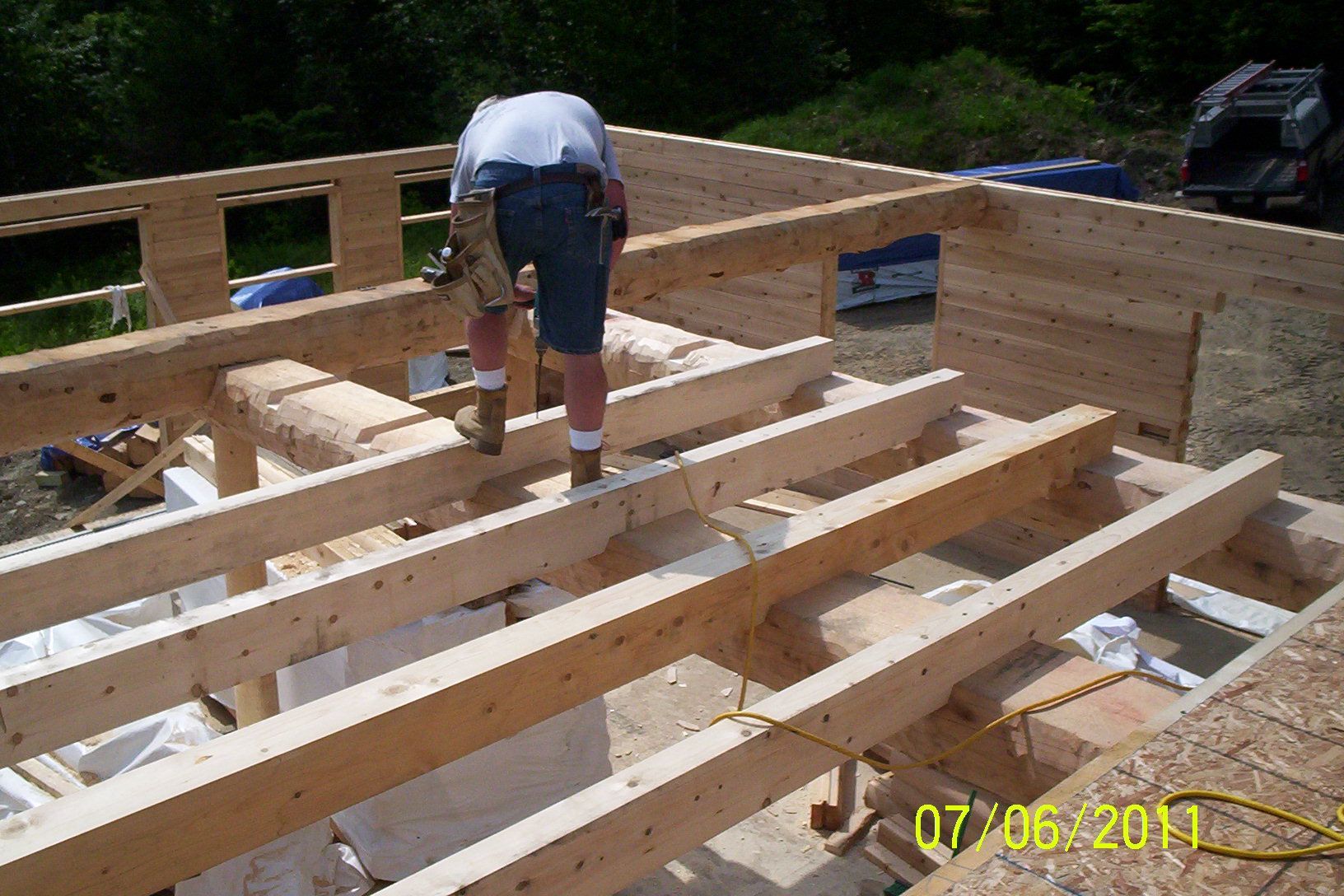 Worker on wooden deck frame construction site, bending over exposed joists outdoors.