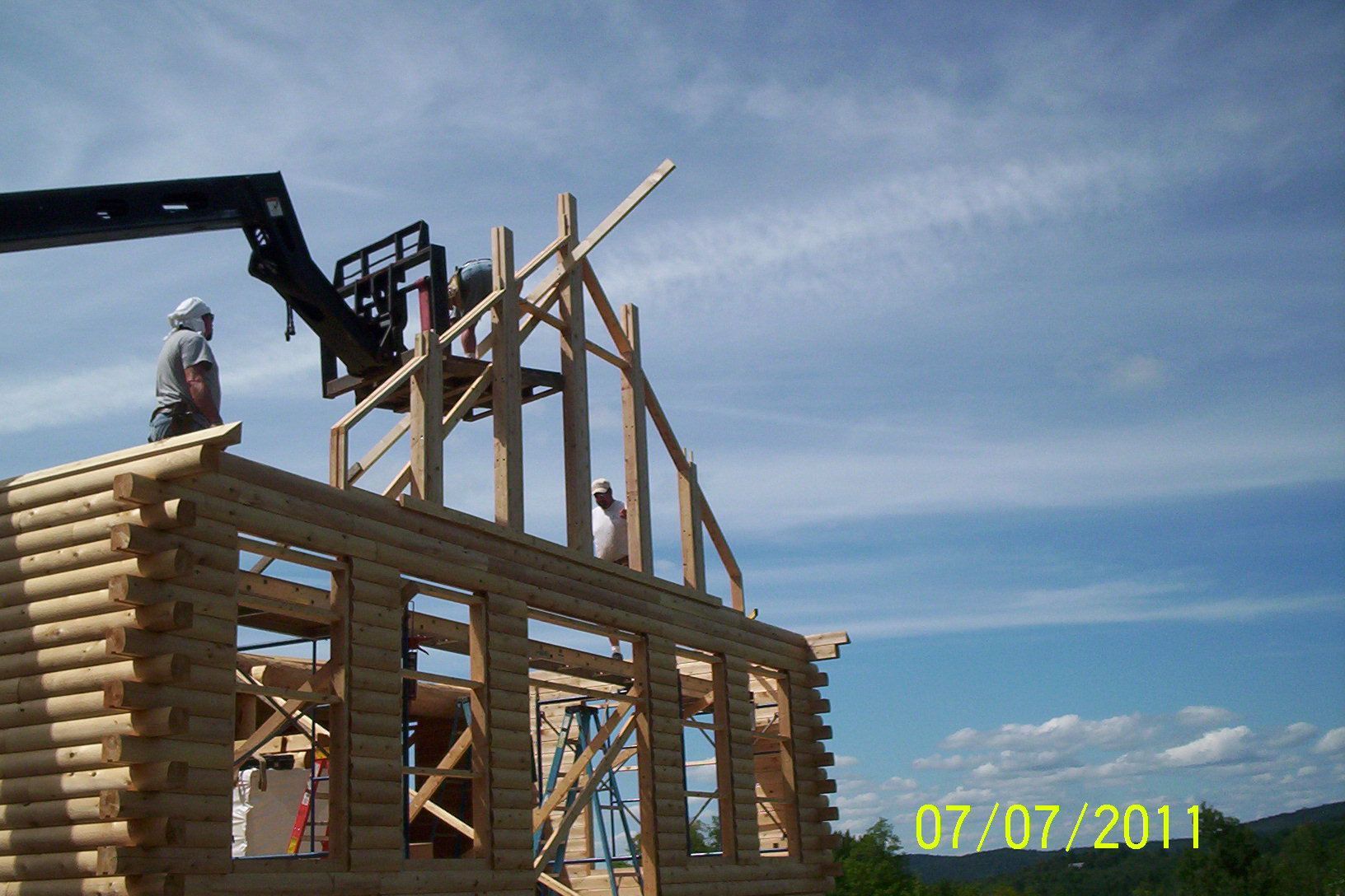 Log cabin under construction with a worker on the roof and a crane lifting framing beams against a blue sky
