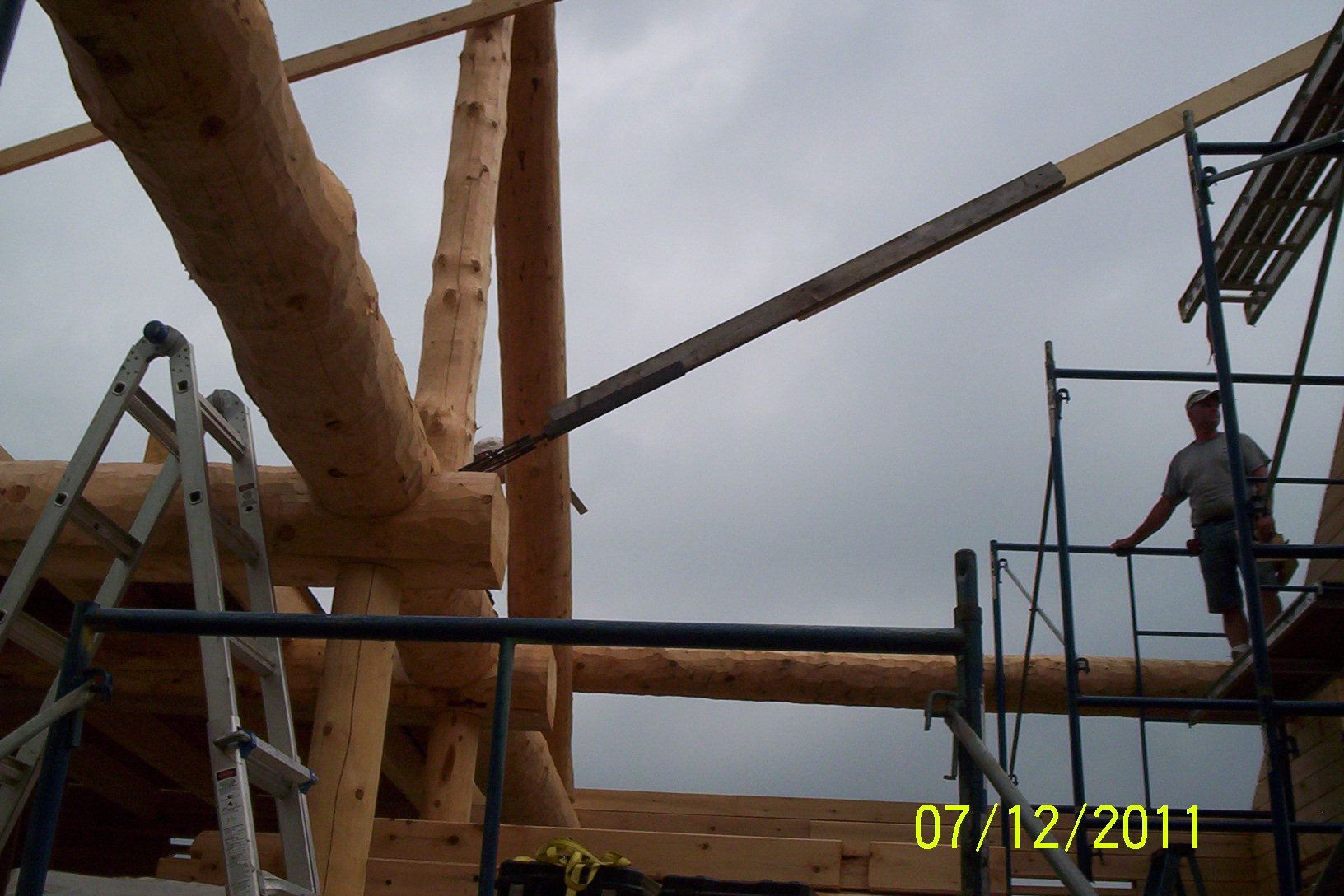 Construction site with large wooden beams, metal scaffolding, and a worker standing on a platform.
