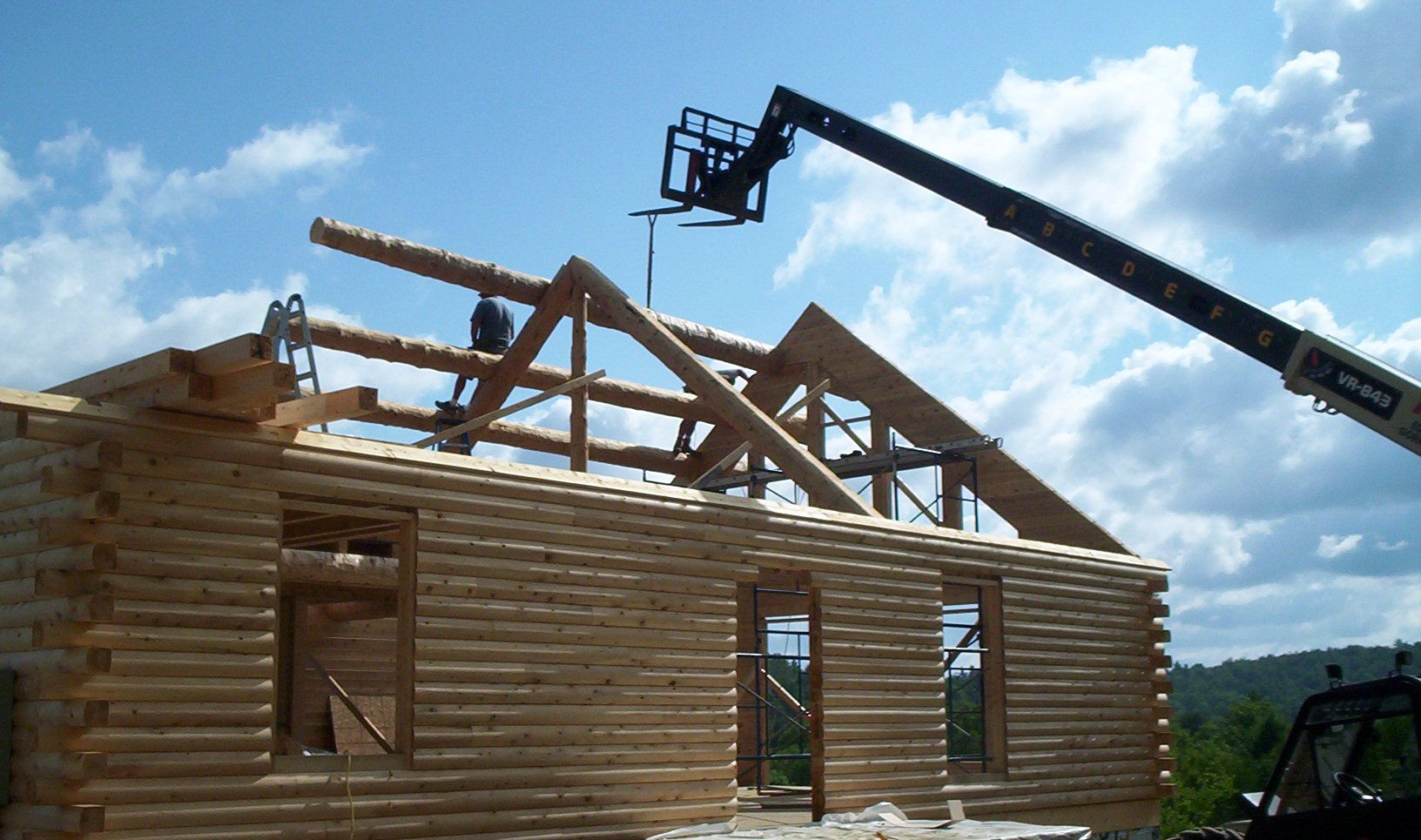 Partially built log cabin with roof beams being lifted by a crane under a blue sky