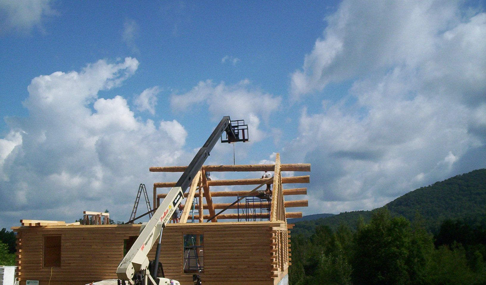 Crane lifting lumber beams onto a wooden house under construction against a blue sky