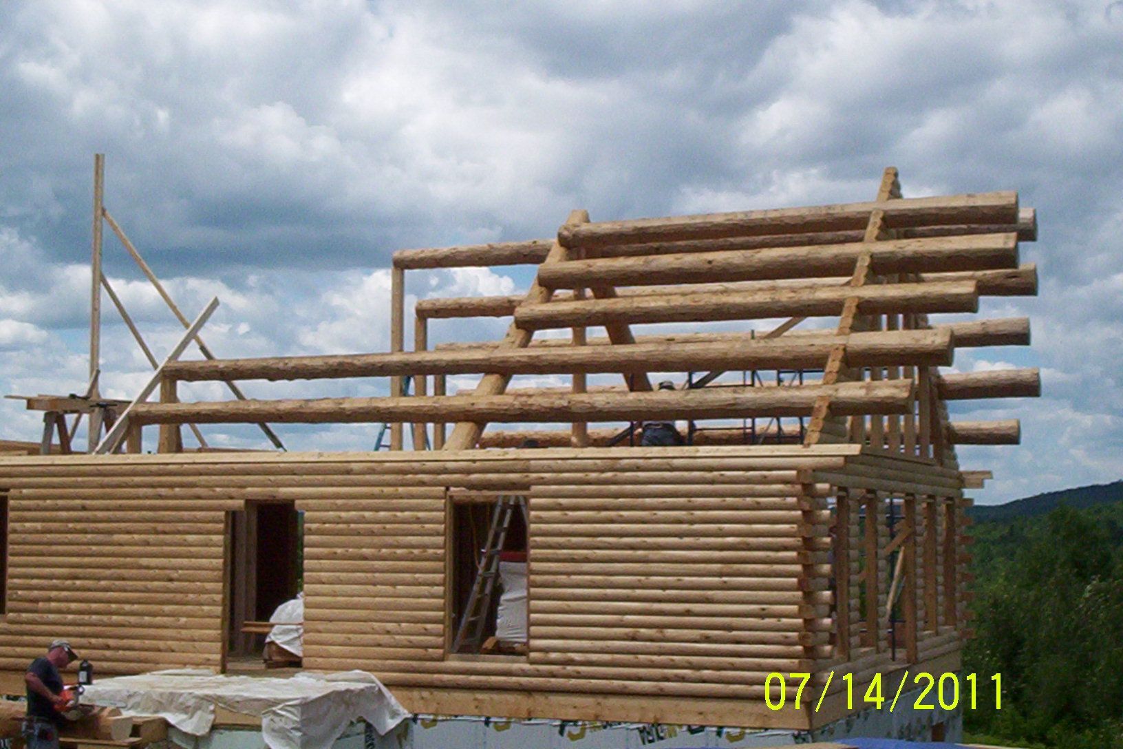 Partially built log cabin with wooden roof framing under a cloudy sky