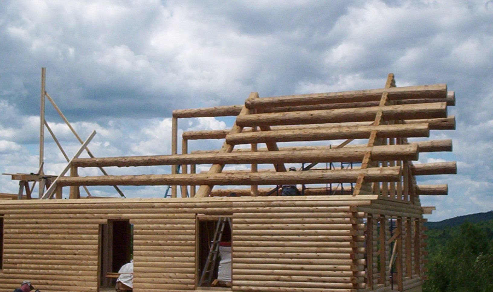 Log cabin under construction with stacked logs and roof beams against a cloudy sky