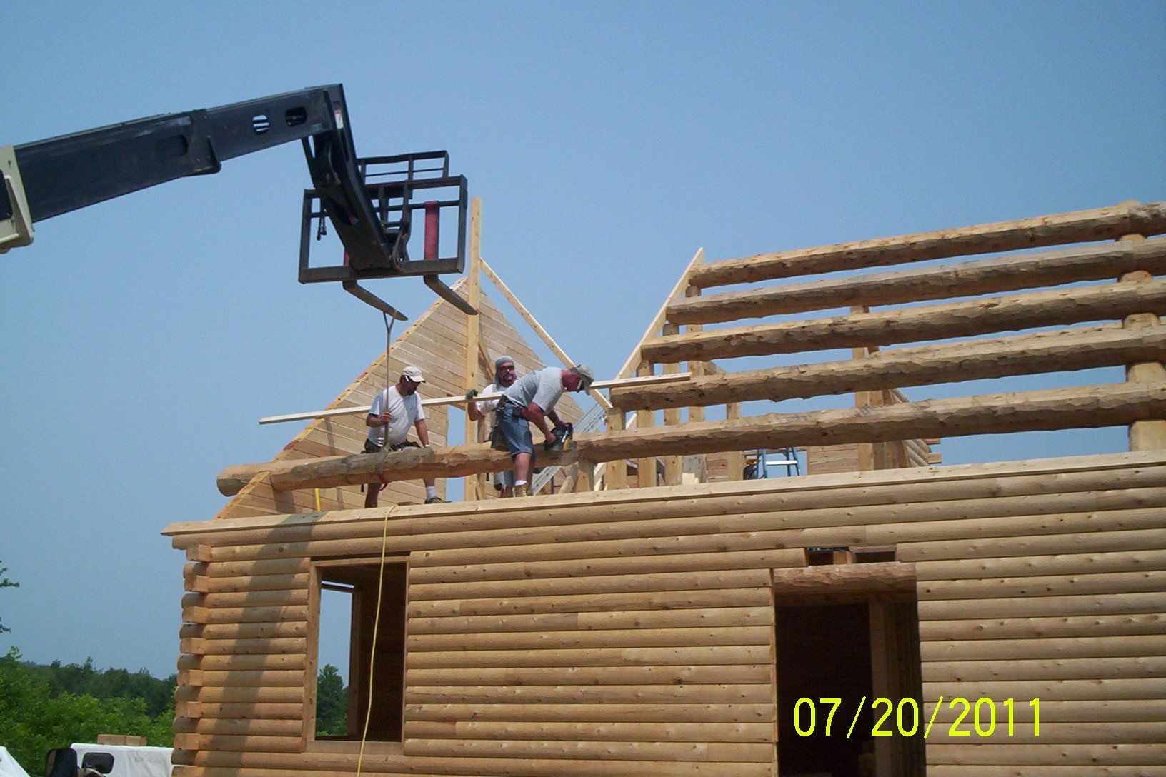 Workers framing a wooden house roof under a crane, with blue sky overhead.