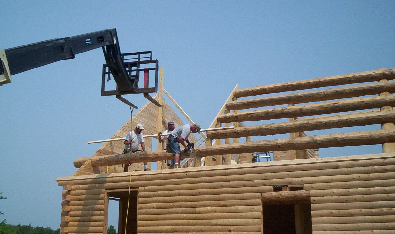 Workers assembling a log cabin roof frame beside a crane under a clear blue sky