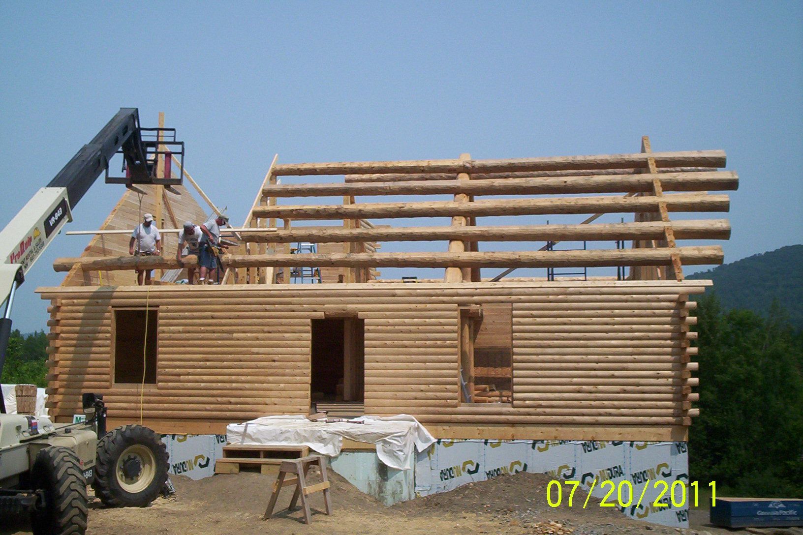 Workers framing a log cabin with a crane, open roof beams, and a tractor in a rural setting