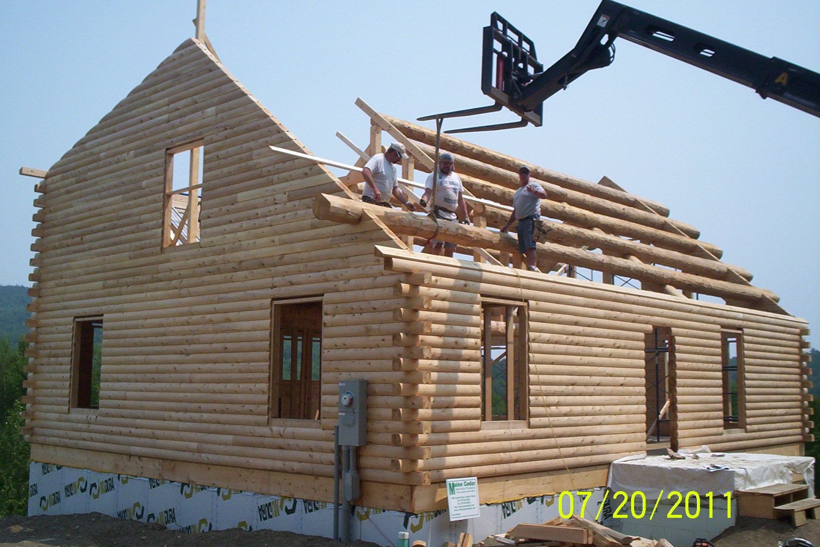 Workers framing a log house roof with a crane, date stamp visible