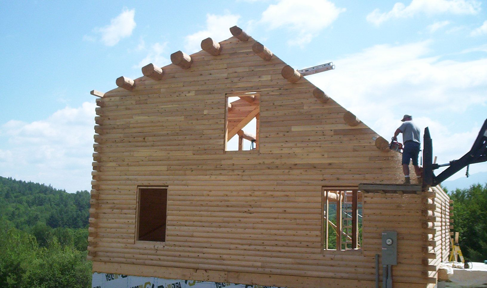 Two people work on a wooden cabin under construction on a grassy hillside.