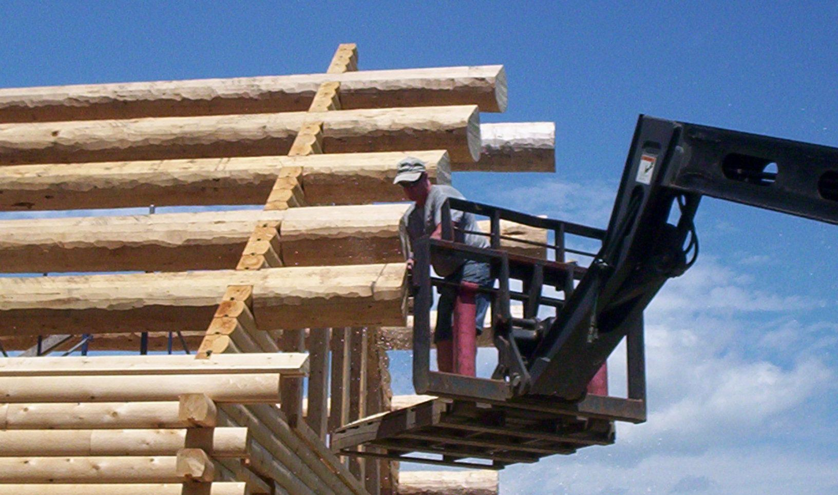 Worker on a lift assembling stacked wooden logs at a construction site under a clear blue sky