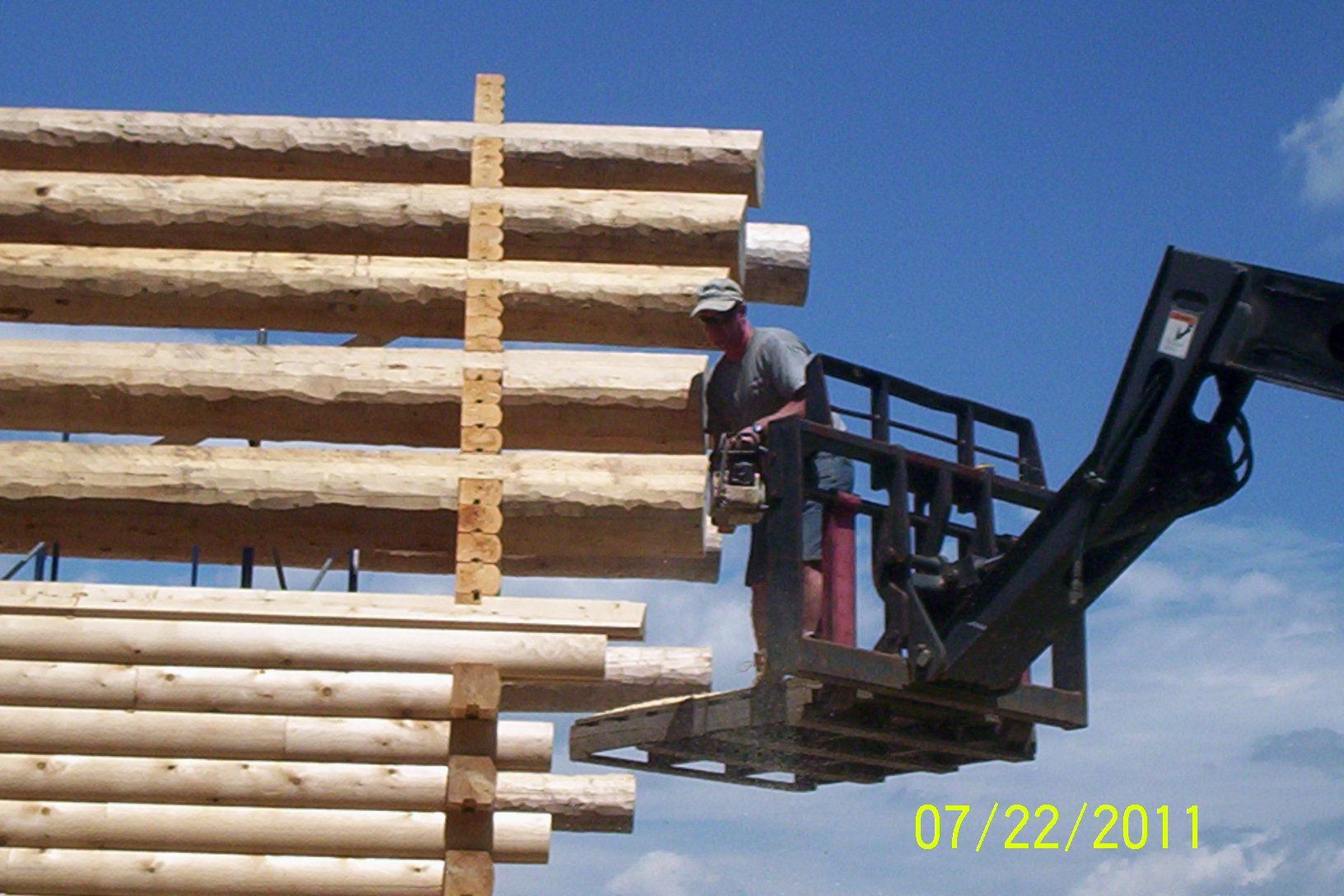 Worker on forklift lifting stacked logs against a blue sky.