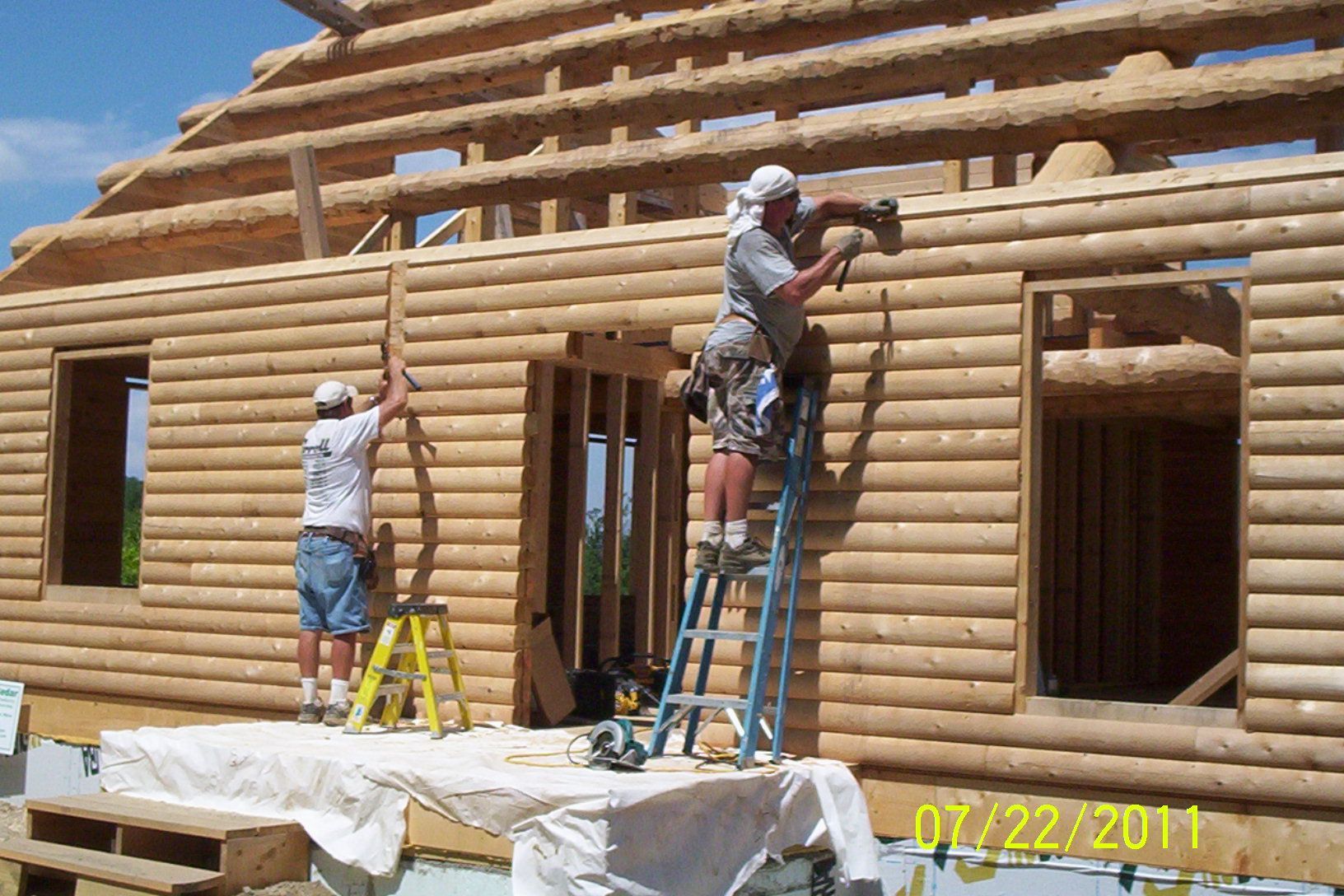 Two workers installing logs on a timber house frame, one on a ladder and one on scaffolding