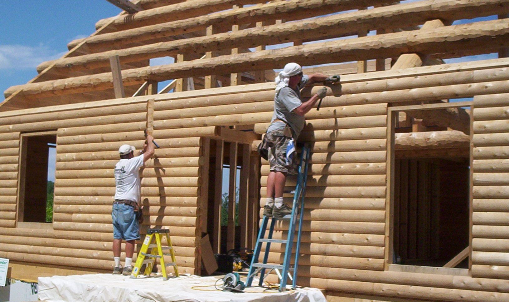 Workers building a log cabin exterior, using ladders and tools on the wooden walls.