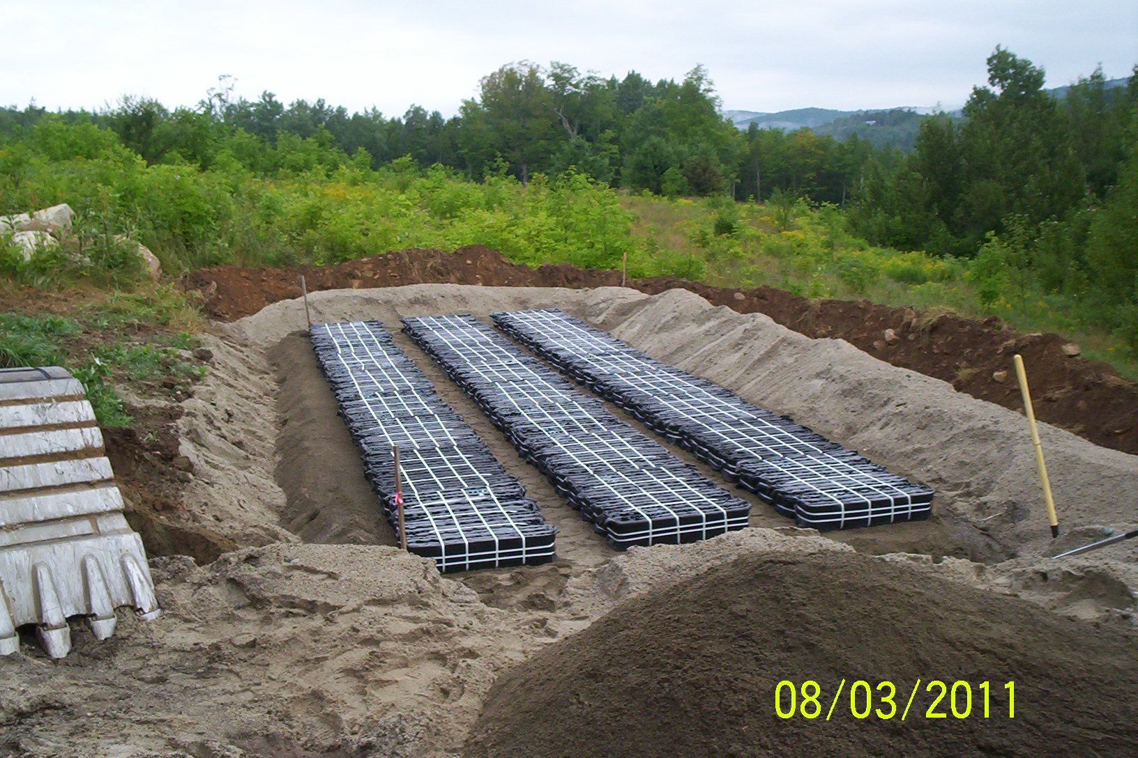 Rectangular excavation filled with rows of metal frame panels in a sandy outdoor site, trees in background