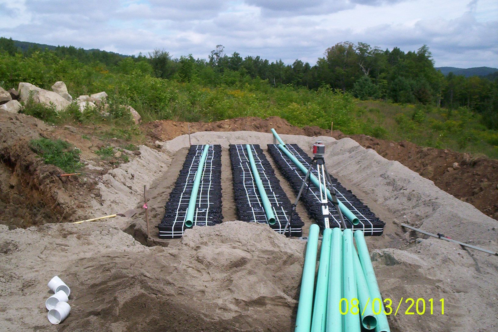 Trenches with black pipes and green tubing laid in sand at a rural construction site, with trees beyond.