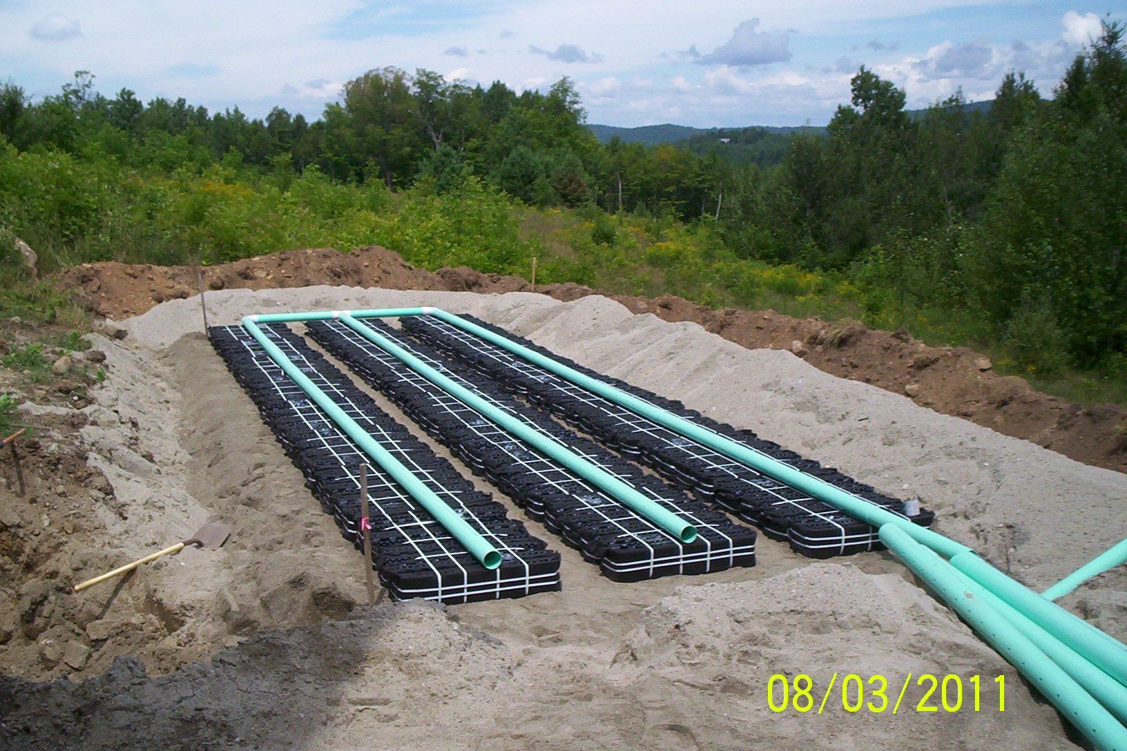 Trenches with black and blue pipes laid in gravel at a construction site, with trees and hills beyond.