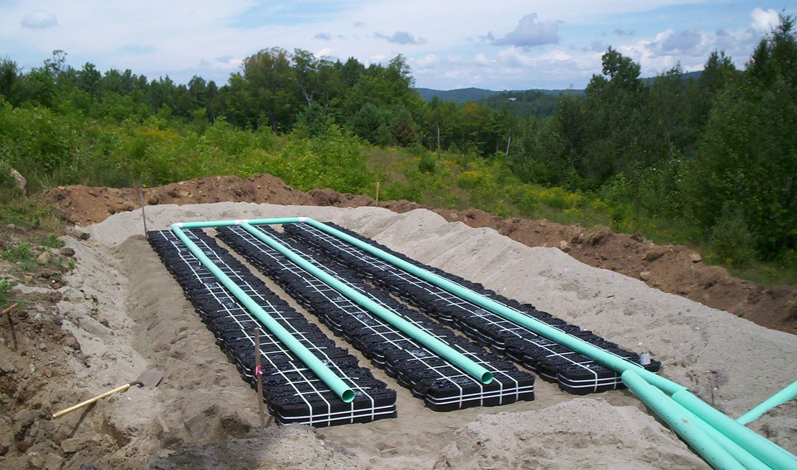 Rows of black pipes and turquoise conduits laid in a gravel trench at an outdoor construction site.