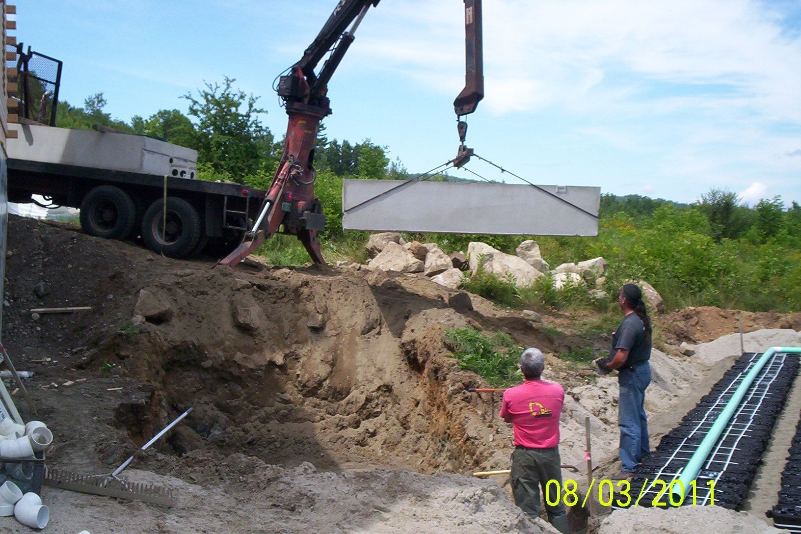 Construction workers using a crane to lower a concrete slab into a dirt excavation beside a truck