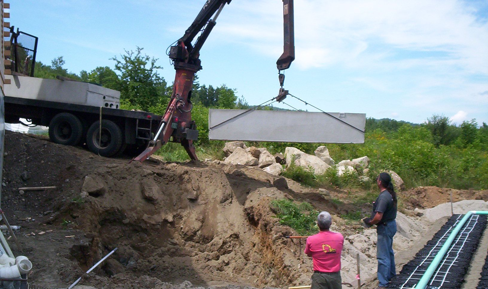 Crane lowering a large concrete slab into an excavation while workers watch from the edge.